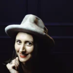 Woman wearing gray Memento fedora hat, front view, dark background, smiling expression