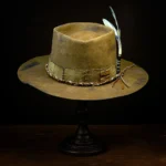 Front view of brown western nomad hat with braided band and feather on wooden stand
