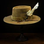 Side view of brown western nomad hat with feather, cords, and playing card accent on wooden stand